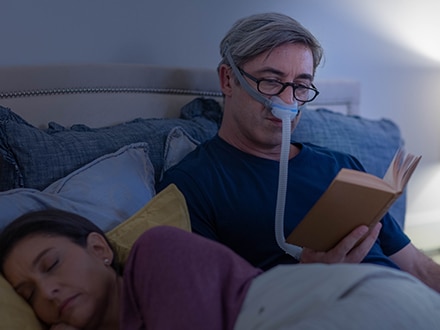 En mand med briller læser i sengen, mens han bærer en ResMed AirFit N30 nasal minimalistisk maske, mens hans kone sover ved siden af ham.