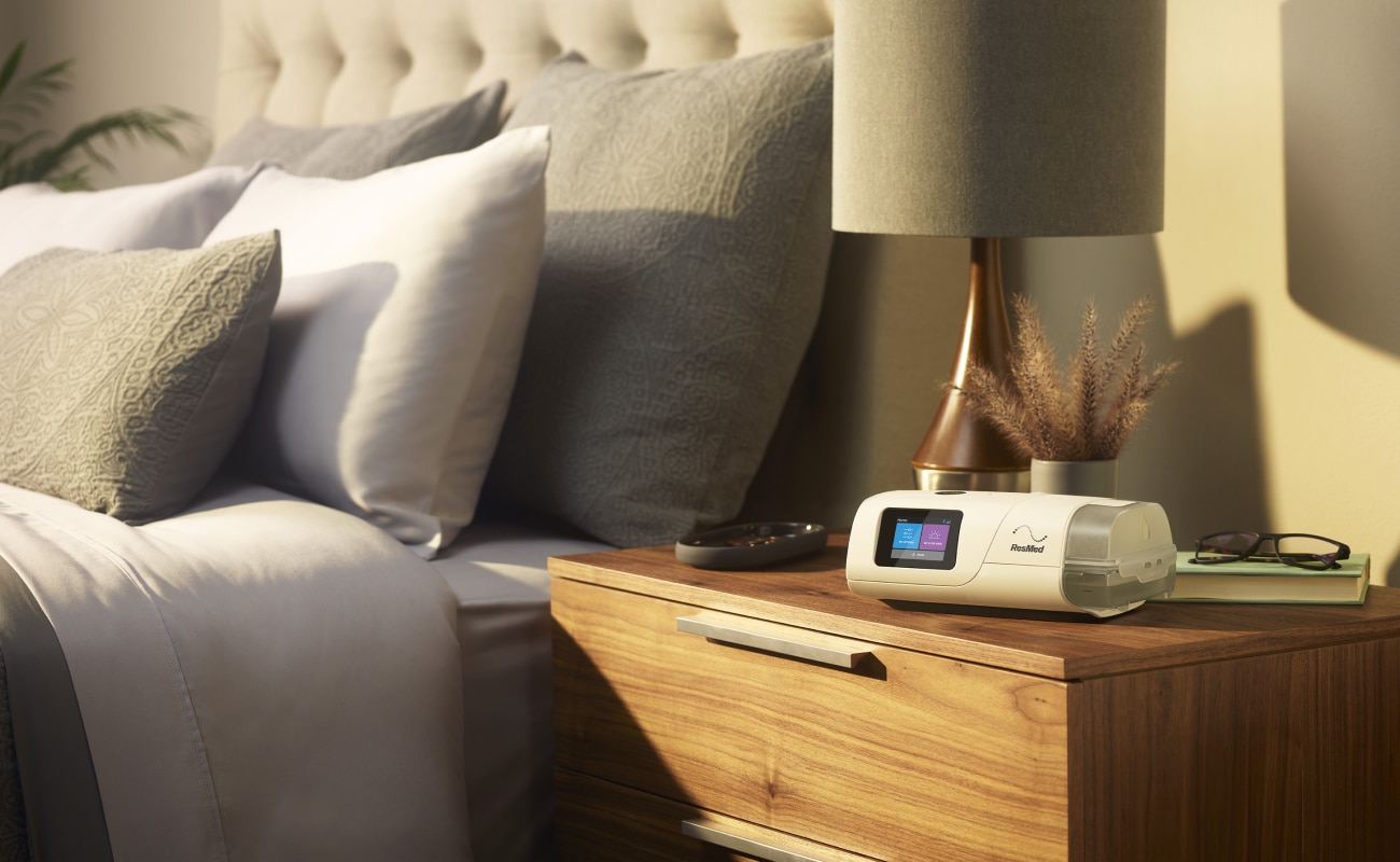 Et AirCurve 11 ASV PaceWave adaptivt servo-ventilationsapparat på et natbord sammen med en lampe, læsebriller og en bog.