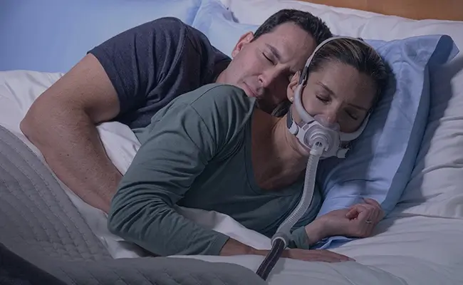 Et par, der sover tæt sammen i sengen, og hvor kvinden har en AirFit F40 CPAP-maske på.